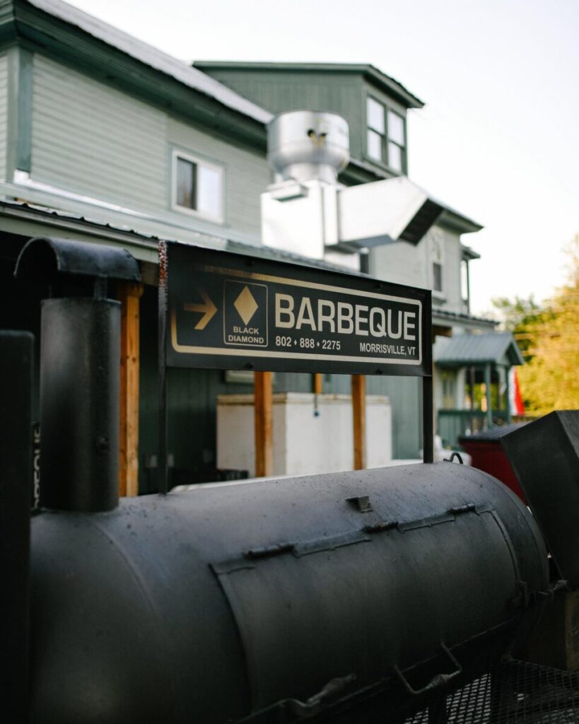 A barbeque smoker sits outside a building with a sign reading "Barbeque, Morrisville, VT, 802-888-2275.