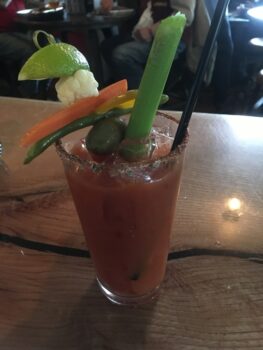 A Bloody Mary cocktail garnished with celery, lime, pickled vegetables, and a spiced rim, on a wooden table.
