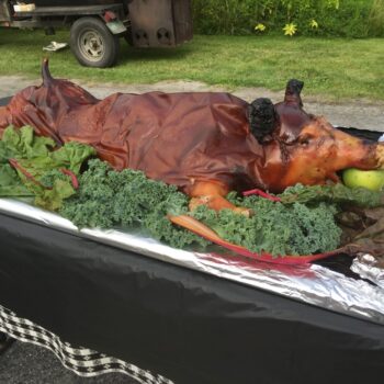 A whole roasted pig with an apple in its mouth is displayed on a table surrounded by leafy greens outdoors.