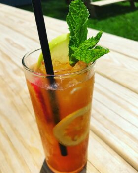 Iced tea with lemon, apple slice, mint garnish, and a black straw on a wooden outdoor table in sunlight.