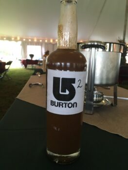 A brown liquid in a glass bottle labeled "BURTON" sits on a table under a tent with catering equipment in the background.