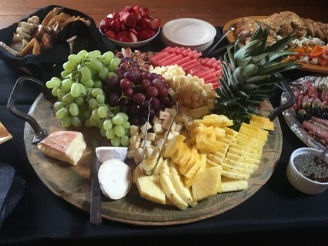 A wooden platter with assorted cheeses, grapes, pineapple slices, watermelon, strawberries, and bread.